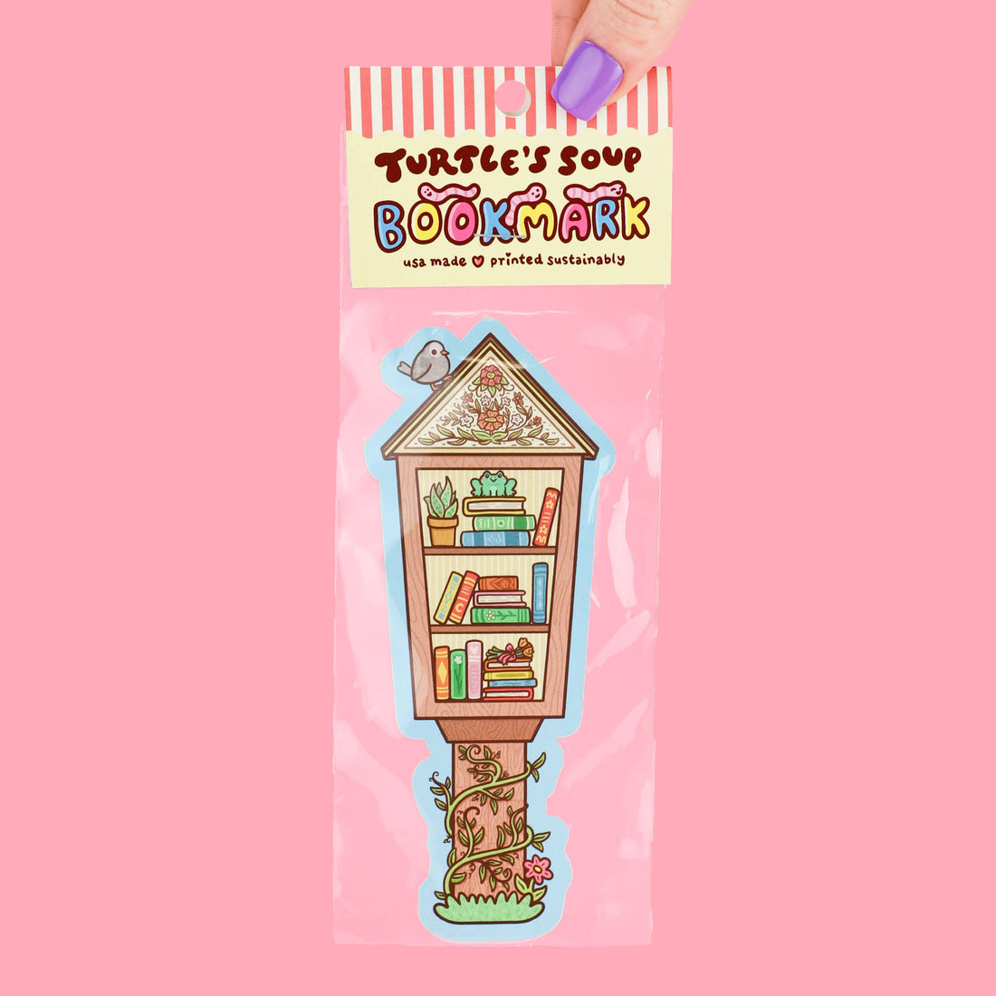 Packaged illustrated bookmark shaped like a tiny wooden bookshelf on stilts with colorful books, plants, flowers, and a small bird perched on the roof. The packaging reads “Turtle’s Soup Bookmark” and “usa made ♥ printed sustainably” against a pastel pink background while a hand with purple nail polish holds the top of the package.