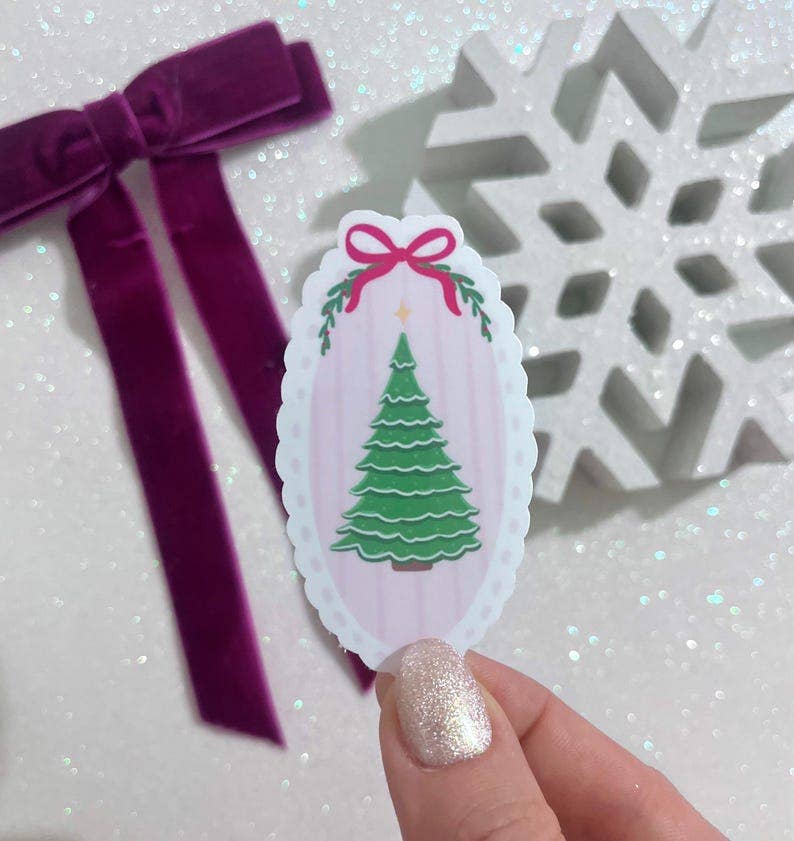 A hand with glittery nail polish holds a scalloped oval sticker featuring a green Christmas tree on a pale pink striped background with a pink bow. A purple velvet bow and a white glittery snowflake ornament are blurred in the background.