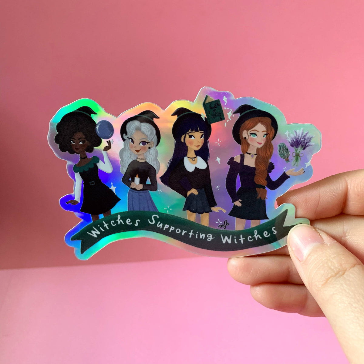 A hand holding a die-cut holographic sticker featuring four cartoon witches in black outfits with the text &