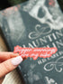Hand holding a pink sticker with text in front of a book titled &