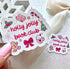 A hand with pink nail polish holds a scalloped white sticker that reads &