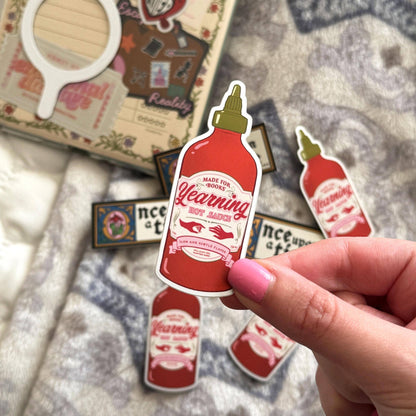 Sticker of a hot sauce bottle with a hand holding it, on a textured surface.