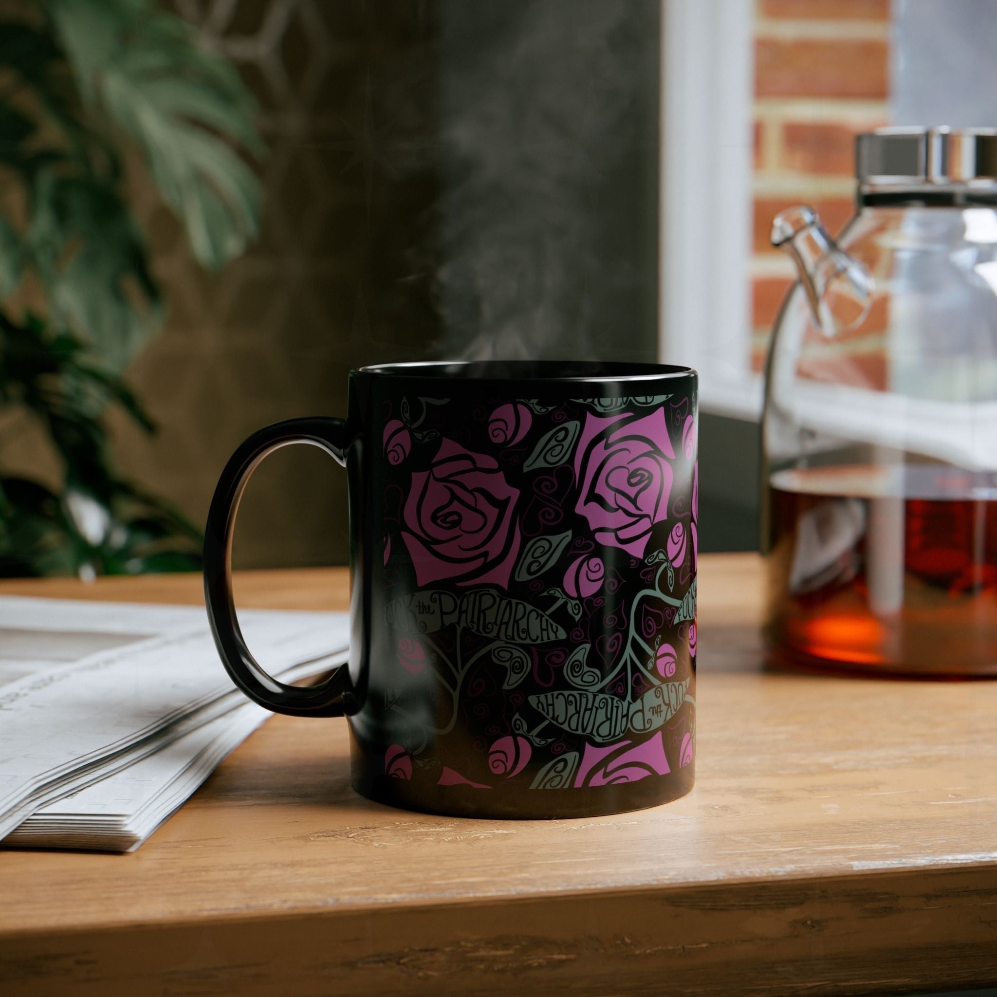 Steaming black feminist coffee mug with fuchsia rose wrap-print pattern on wooden desk beside glass teapot; cozy reading lifestyle scene