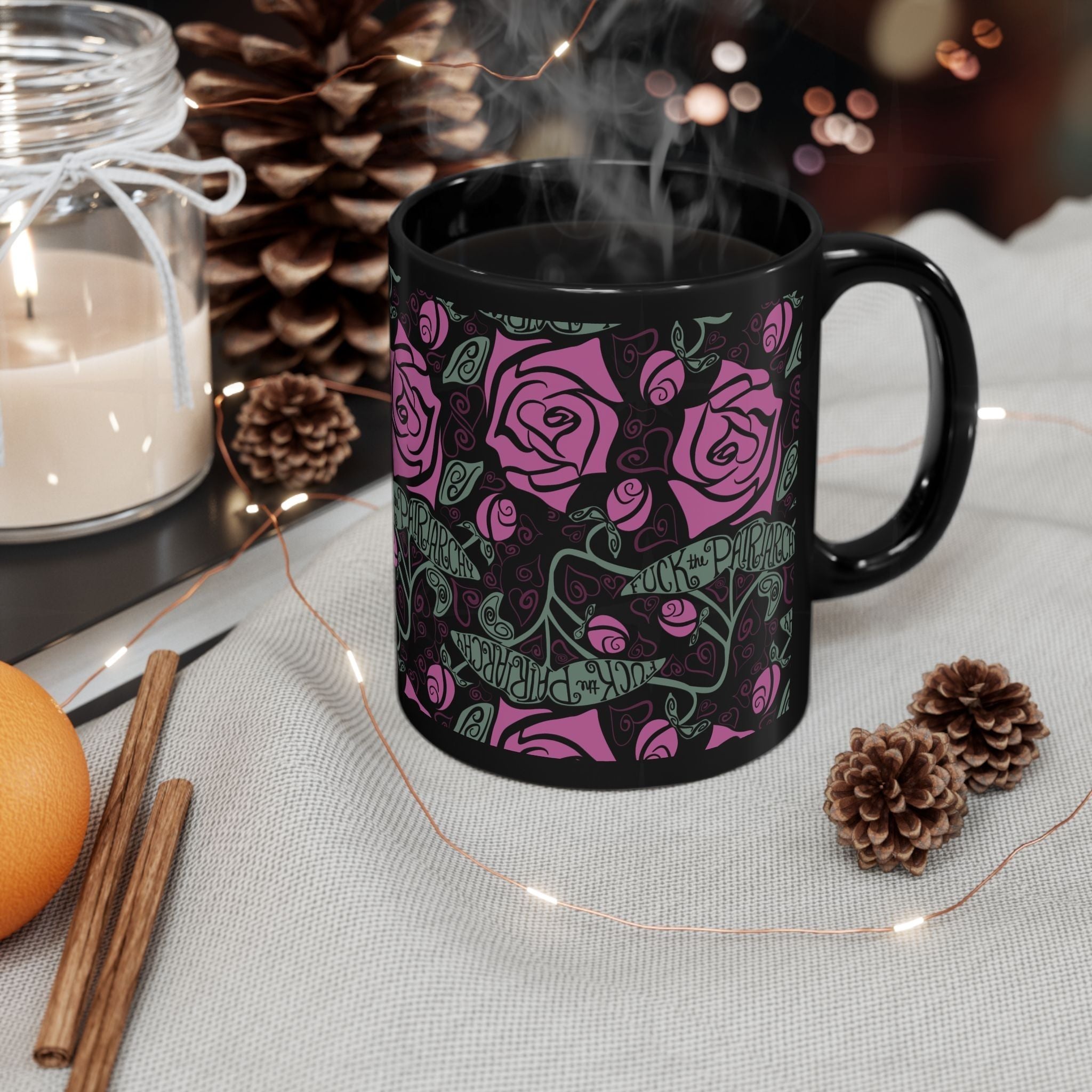 Steaming black ceramic feminist mug with fuchsia rose wrap-print surrounded by pine cones, cinnamon sticks, and candles — cozy holiday gift scene