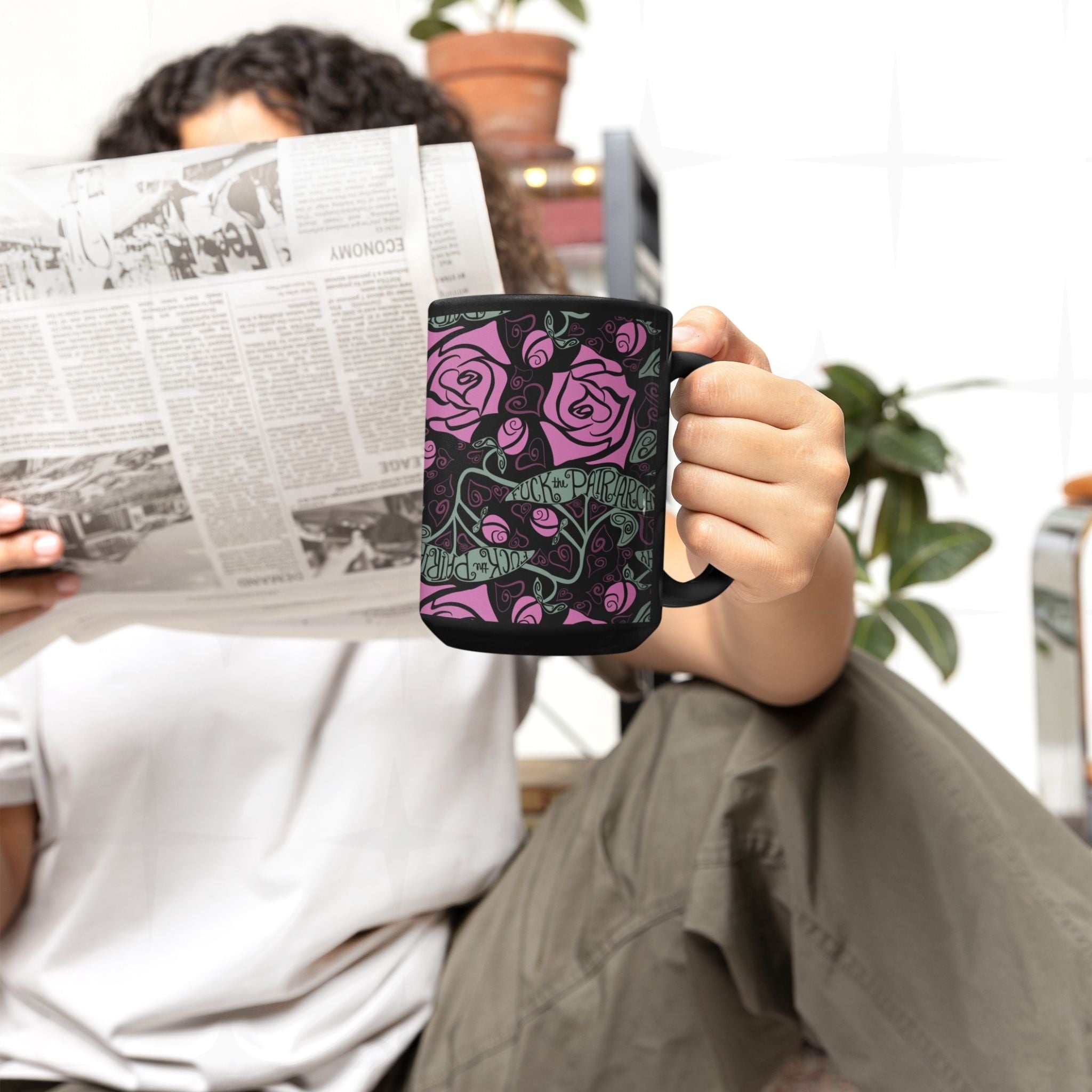 Person holding 15oz black ceramic feminist mug with fuchsia rose wrap-print pattern while reading newspaper; lifestyle scene
