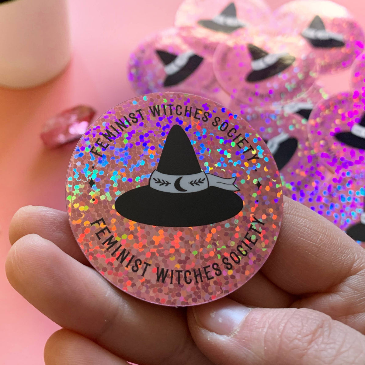 A hand holds a round glittery pink pinback button featuring a black witch hat graphic and the text &