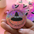 A hand holds a round glittery pink pinback button featuring a black witch hat graphic and the text &
