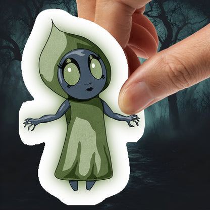 Hand holding the Flatwoods Monster sticker, a gray figure in a green hood with large pale eyes, against dark woods