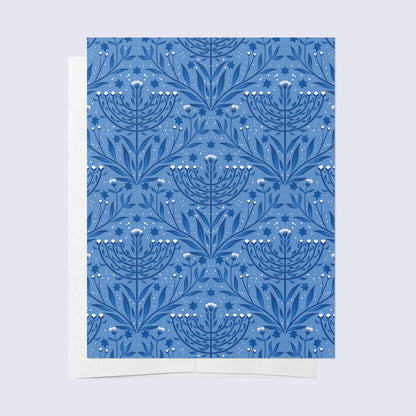 A folded greeting card with a blue background and a white floral Menorah pattern, paired with a bright white envelope.