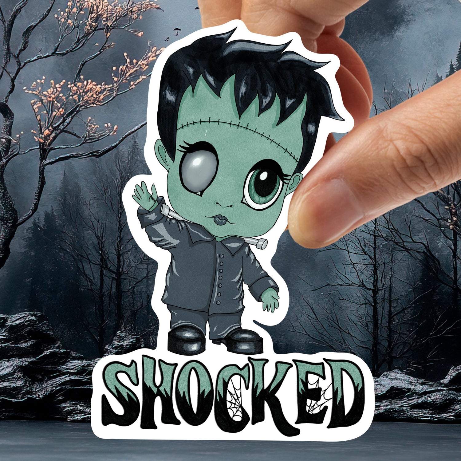 Hand holding the pastel goth Frankenstein sticker against a dark woodland background