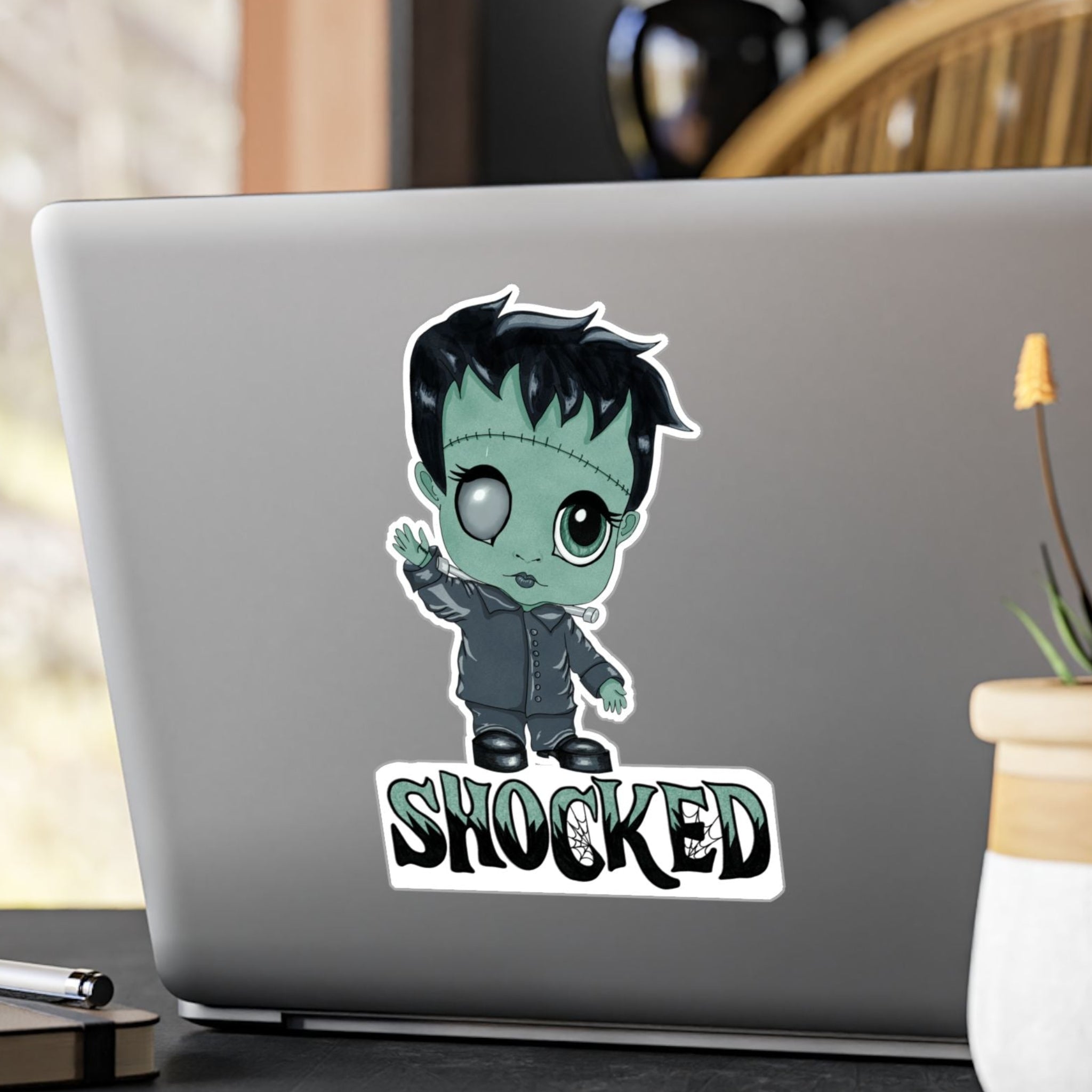 Large chibi Frankenstein sticker with mint-green skin and SHOCKED lettering on the back of a silver laptop