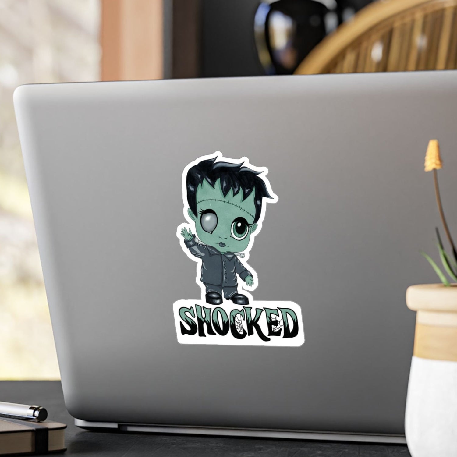 Medium chibi Frankenstein sticker with mint-green skin and SHOCKED lettering on the back of a silver laptop