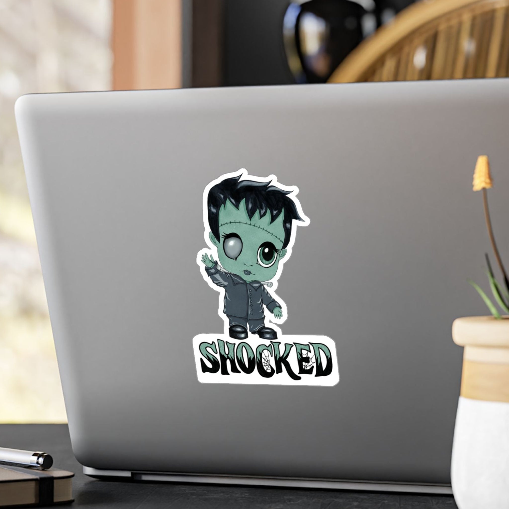 Medium chibi Frankenstein sticker with mint-green skin and SHOCKED lettering on the back of a silver laptop