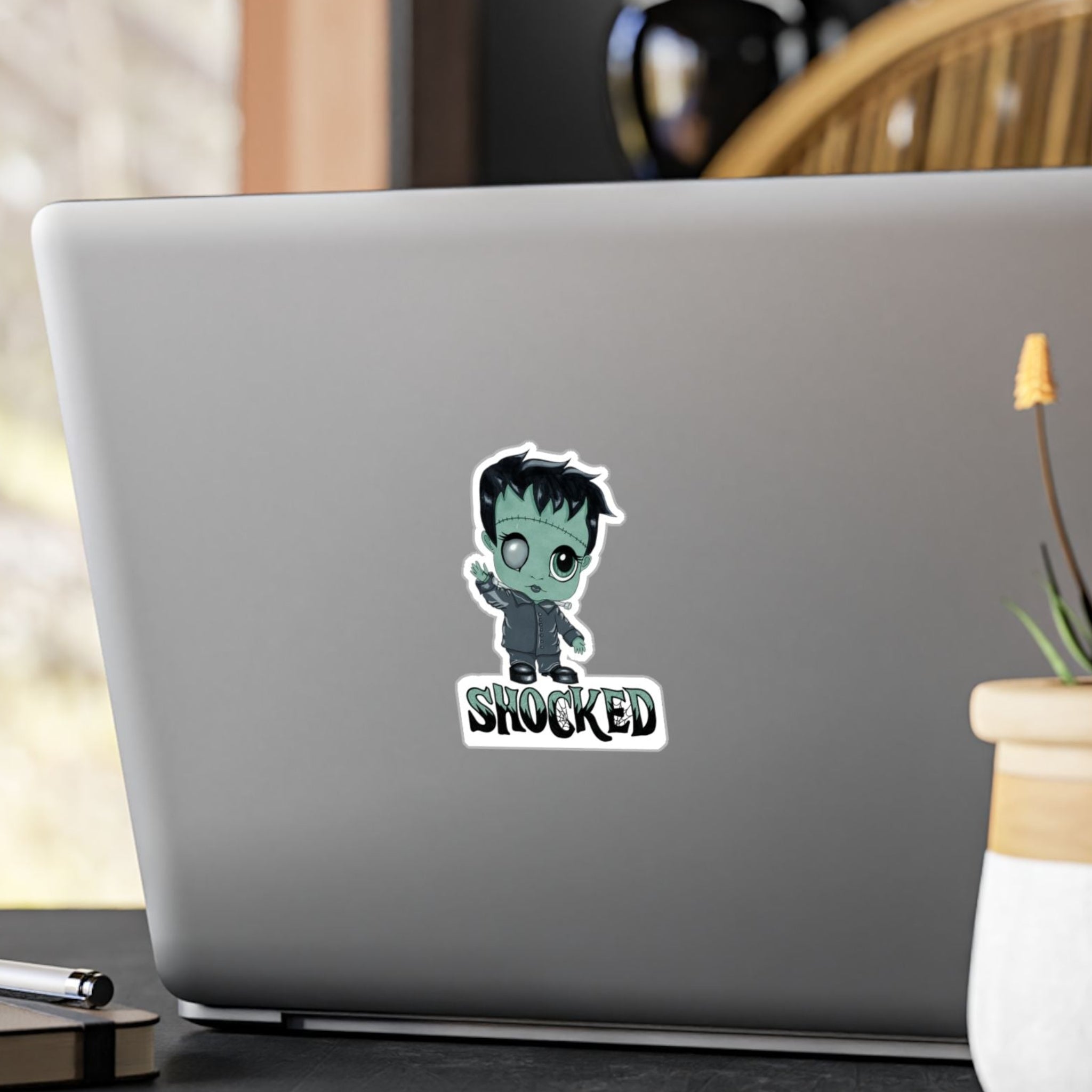 Small chibi Frankenstein sticker with mint-green skin and SHOCKED lettering on the back of a silver laptop