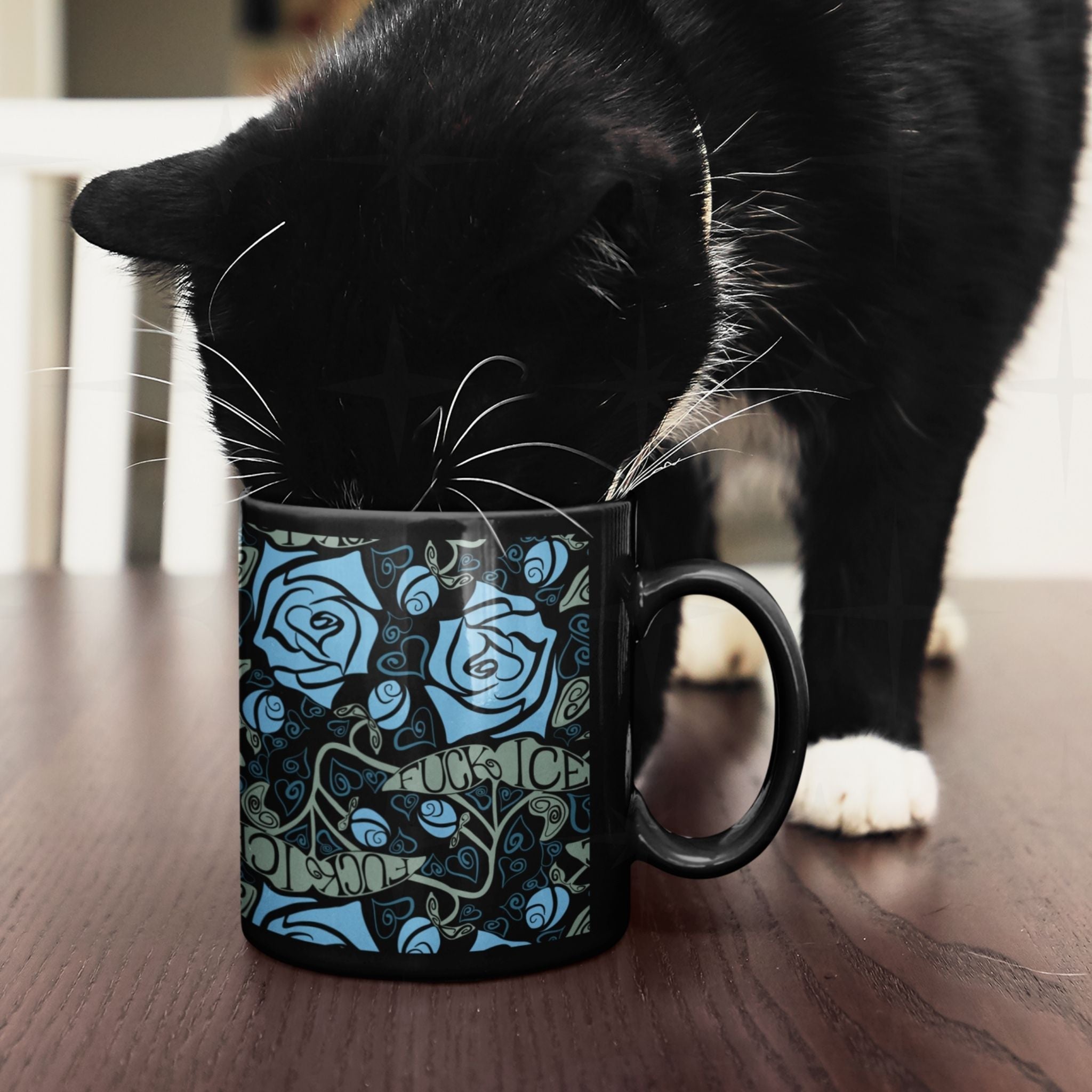 Black cat investigating a dark floral activist coffee mug on a wooden surface, mug shows full botanical rose and hidden fuck ICE leaf message pattern on glossy black ceramic