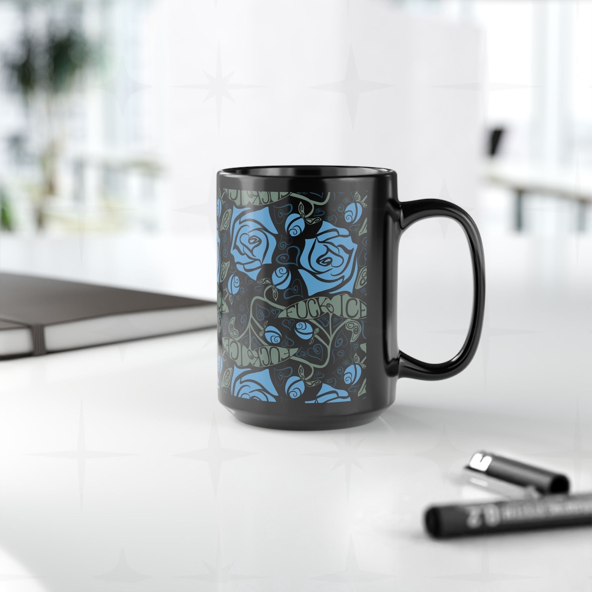 Dark floral hidden message activist mug sitting on a white desk beside a laptop and markers, showing the botanical rose pattern and concealed anti-ICE lettering in sage green on a 15oz glossy black ceramic mug