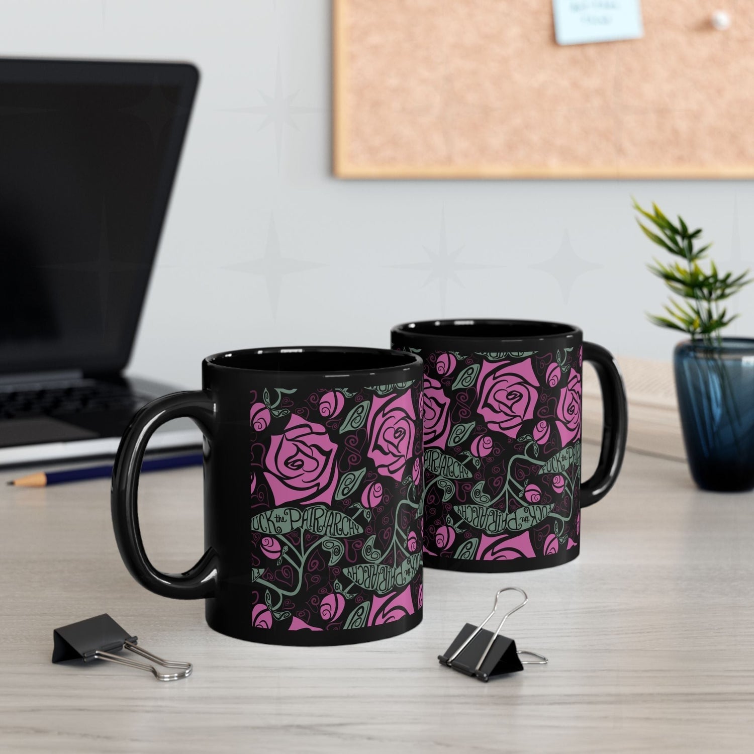 Two black ceramic feminist coffee mugs with fuchsia rose wrap-print on office desk beside laptop and binder clips