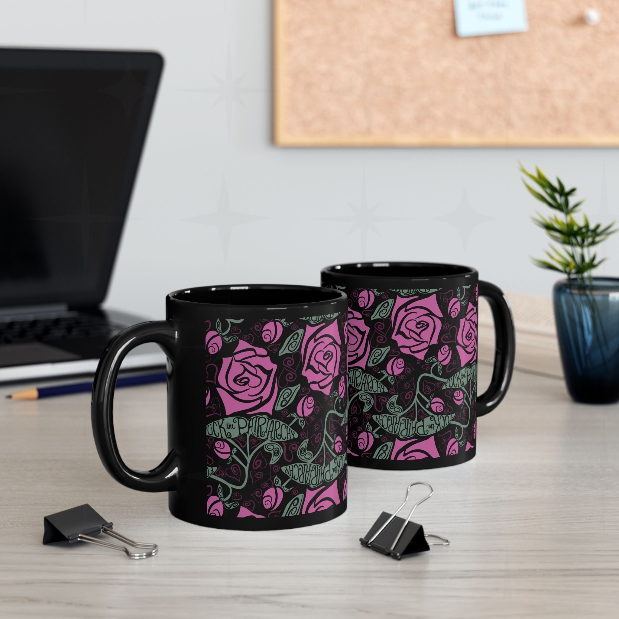 Two black ceramic feminist coffee mugs with fuchsia rose wrap-print on office desk beside laptop and binder clips