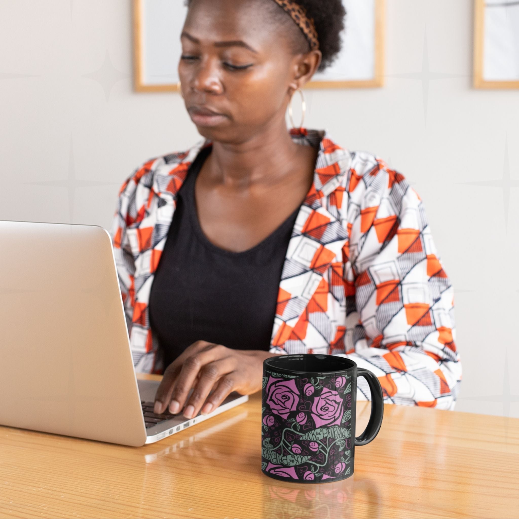 Woman working at laptop with black ceramic feminist coffee mug featuring fuchsia rose and Fuck the Patriarchy pattern on desk beside her