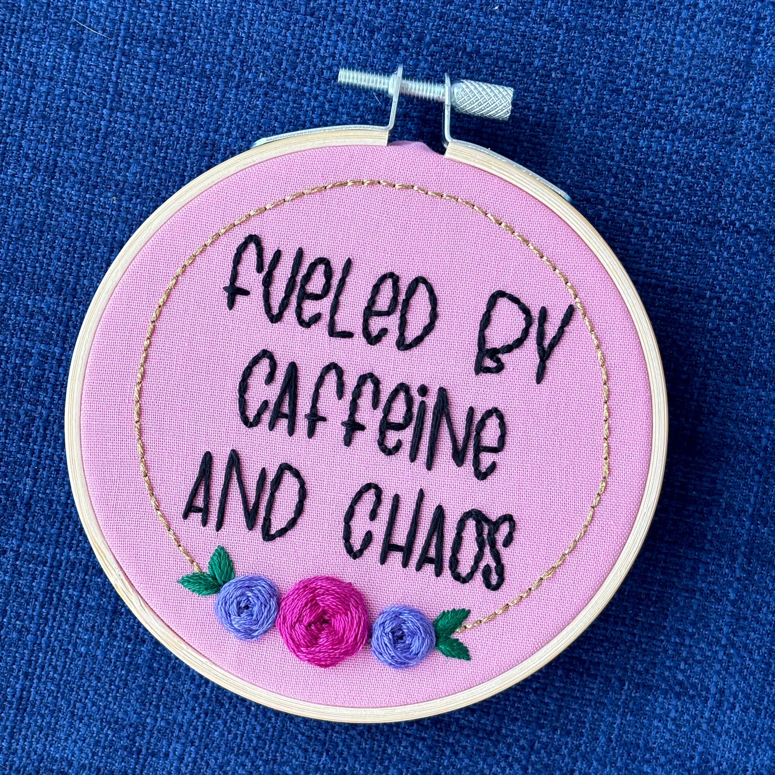 Finished embroidery hoop reads, "fueled by caffeine and chaos". Quote in handwriting print font and hand embroidered in black. Words are centered in a floral frame, which is metallic gold and embellished with cheerful pink and purple roses and dark green leaves sewn with embroidery thread. Fabric is pink cotton. Framed in light wood embroidery hoop as wall art.