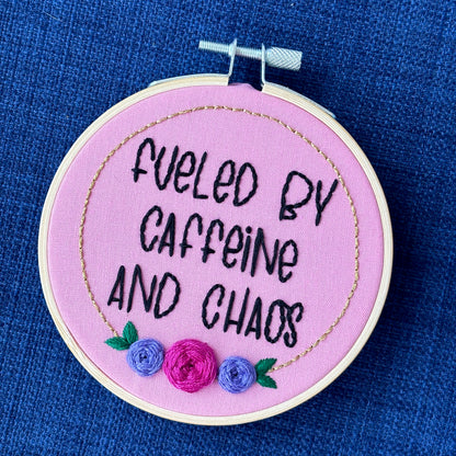 Finished embroidery hoop reads, "fueled by caffeine and chaos". Quote in handwriting print font and hand embroidered in black. Words are centered in a floral frame, which is metallic gold and embellished with cheerful pink and purple roses and dark green leaves sewn with embroidery thread. Fabric is pink cotton. Framed in light wood embroidery hoop as wall art.