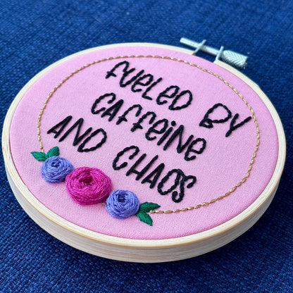Finished embroidery hoop reads, "fueled by caffeine and chaos". Quote in handwriting print font and hand embroidered in black. Words are centered in a floral frame, which is metallic gold and embellished with cheerful pink and purple roses and dark green leaves sewn with embroidery thread. Fabric is pink cotton. Framed in light wood embroidery hoop as wall art.