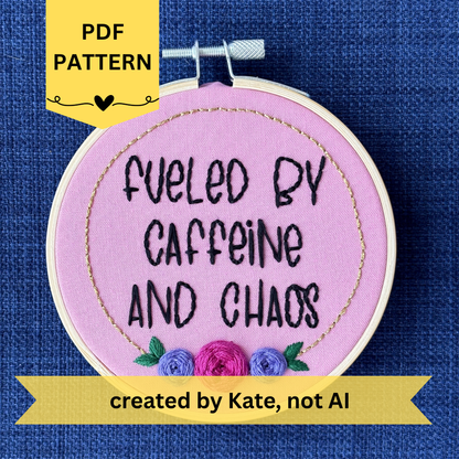 PDF pattern created by Kate, not AI. Finished embroidery reads, "fueled by caffeine and chaos". Quote in handwriting print font and hand embroidered in black. Words are centered in a floral frame, which is metallic gold and embellished with cheerful pink and purple roses and dark green leaves sewn with embroidery thread. Fabric is pink cotton. Framed in light wood embroidery hoop as wall art.