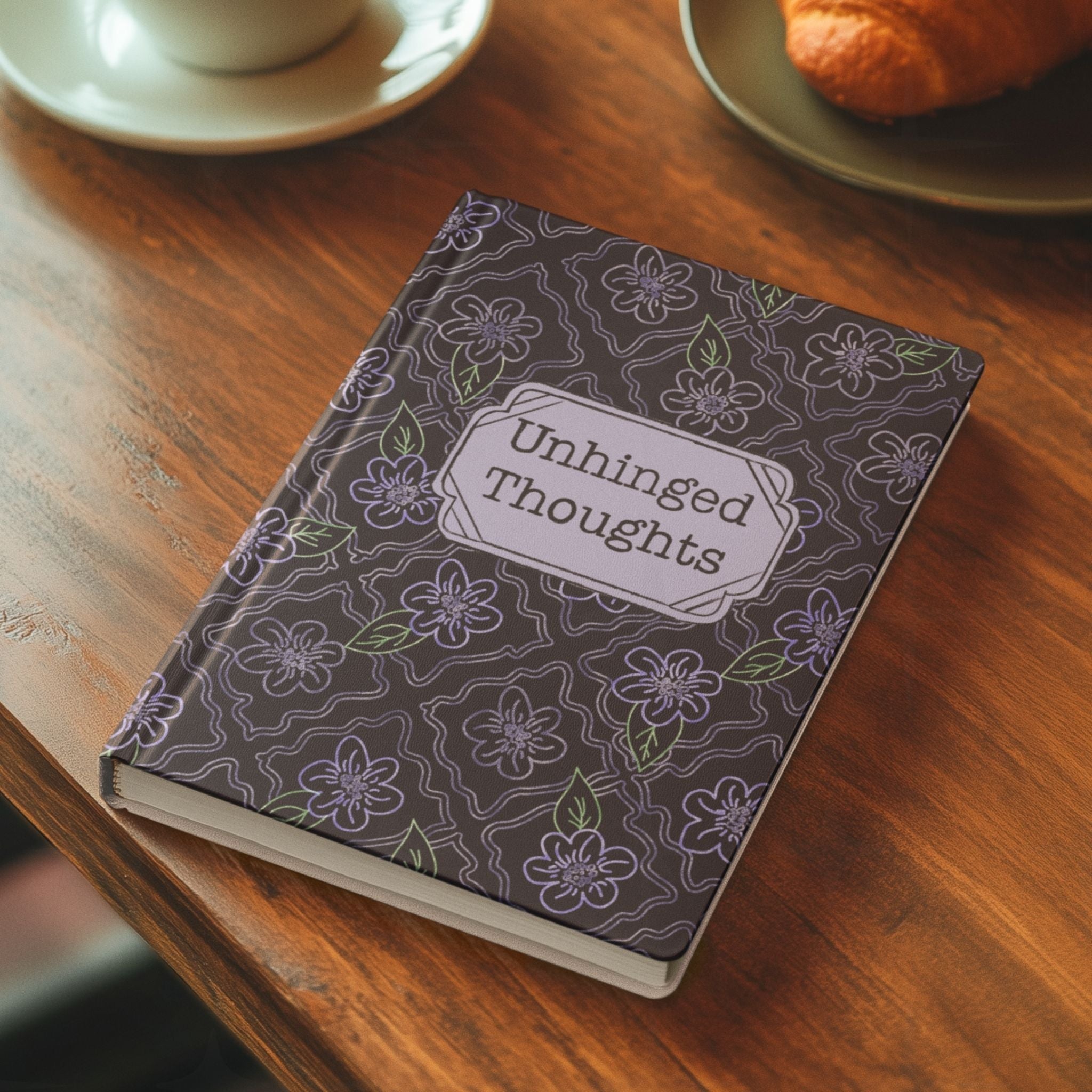 Funny floral hardcover journal on wooden café table beside a cup of coffee and croissant showing Unhinged Thoughts lavender cover design