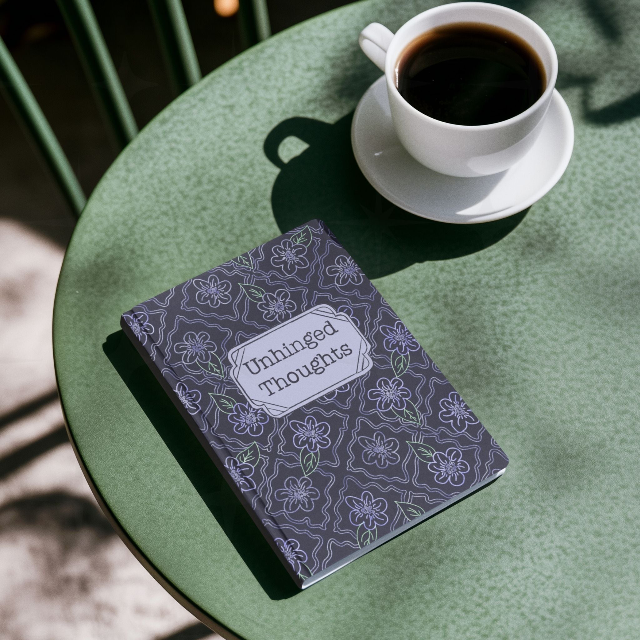 Unhinged Thoughts hardcover journal on green café table beside a white cup of black coffee, overhead view