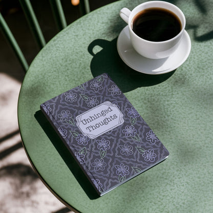 Unhinged Thoughts hardcover journal on green café table beside a white cup of black coffee, overhead view