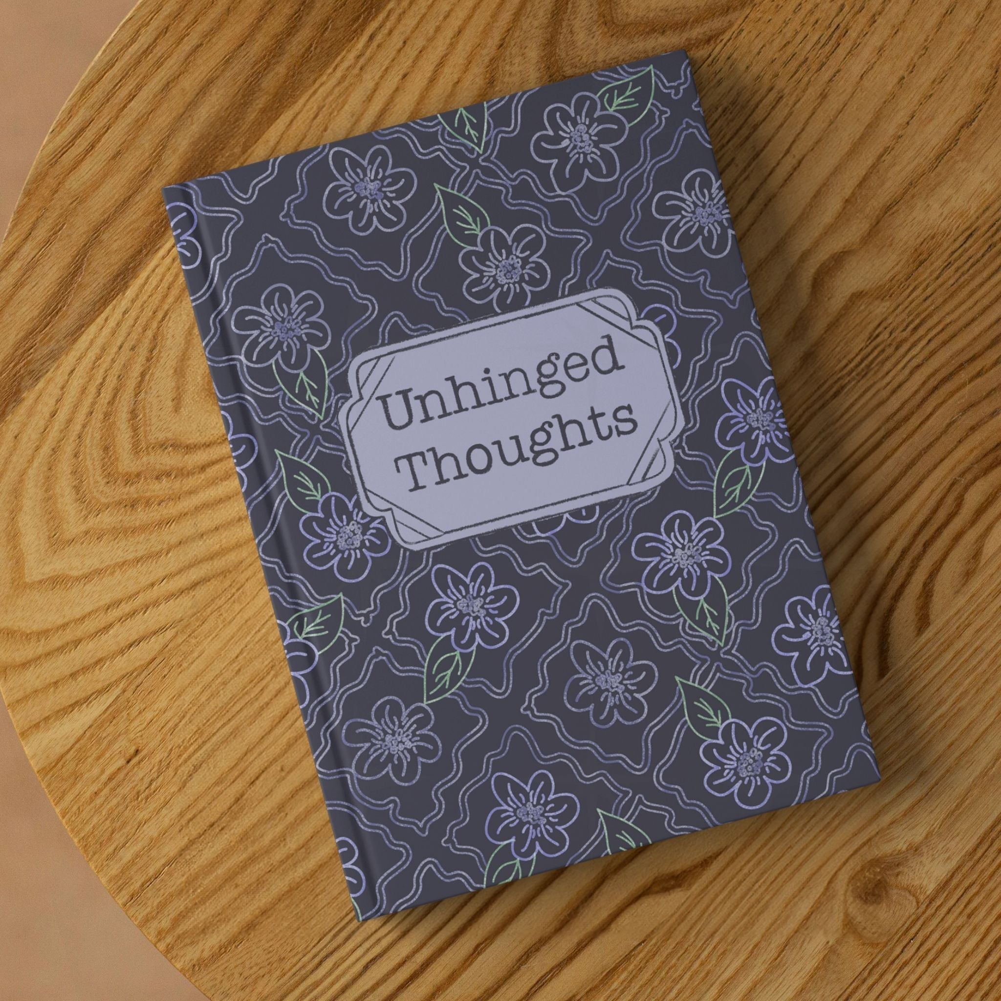 Funny floral hardcover journal with Unhinged Thoughts cover lying on round wooden surface, angled view showing charcoal and lavender ogee pattern