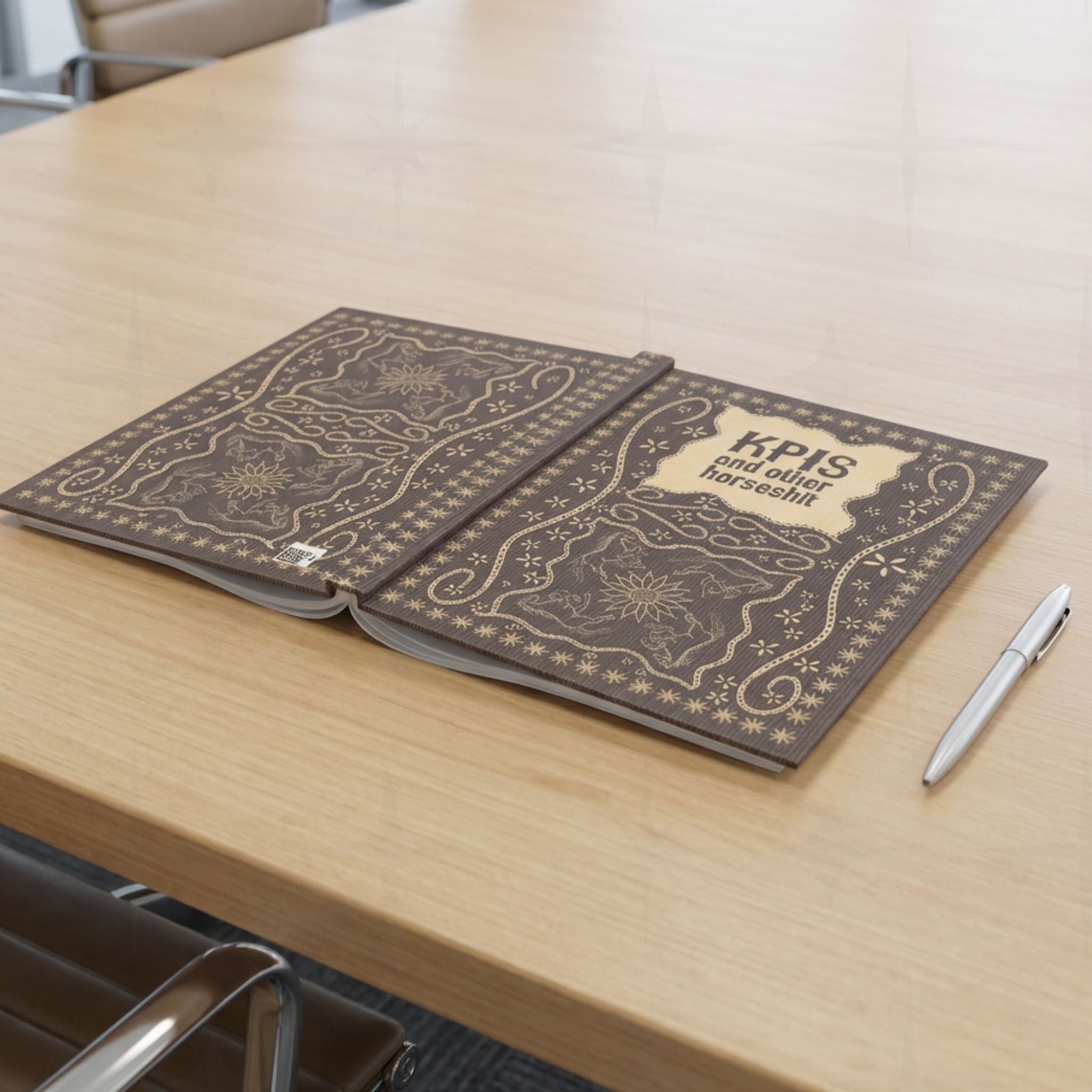 KPI profanity journal lying open flat on conference table beside silver pen showing both covers with vintage equestrian embroidered design