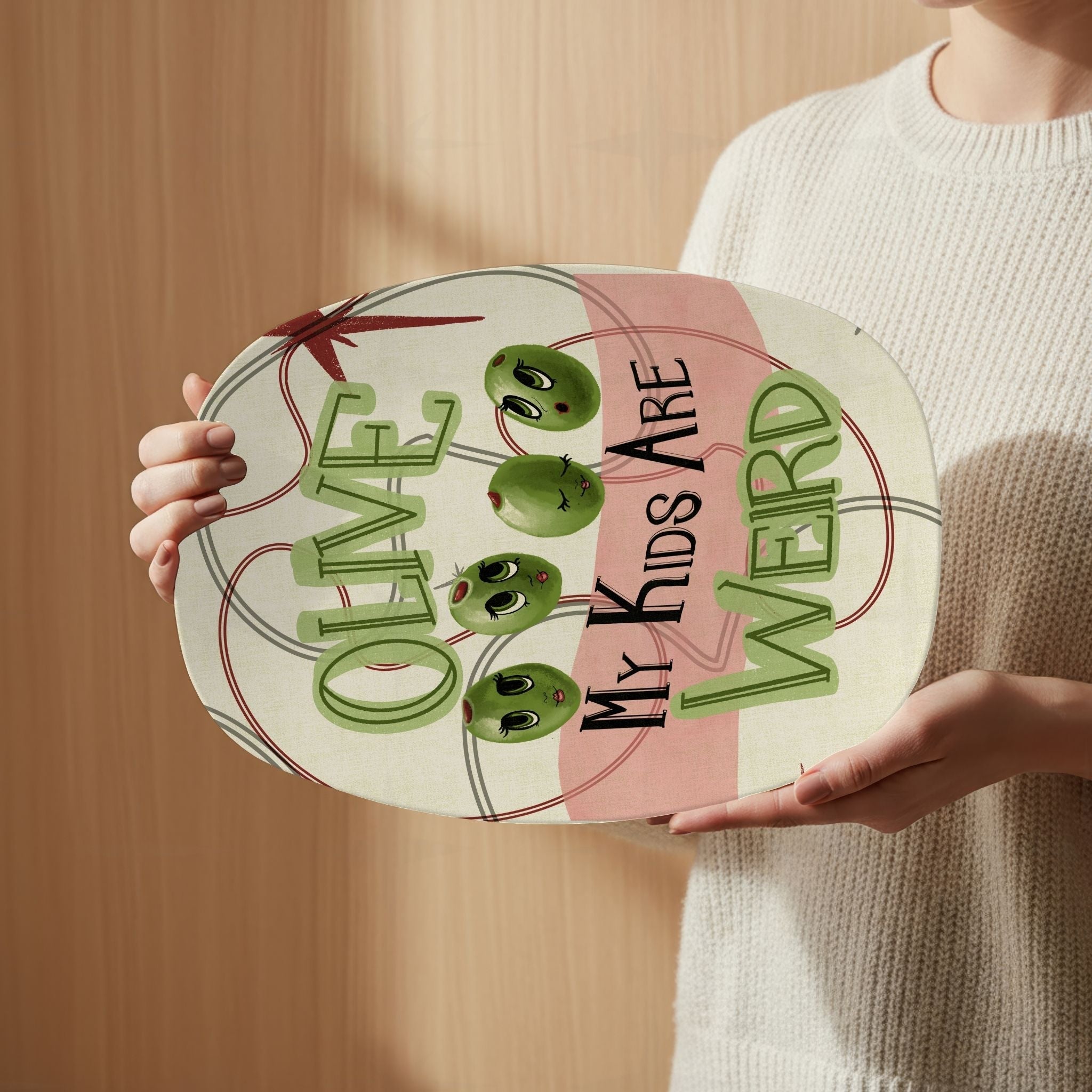 Person in cream sweater holding funny retro serving platter with mid-century olive pun illustration reading Olive My Kids Are Weird against warm wood background