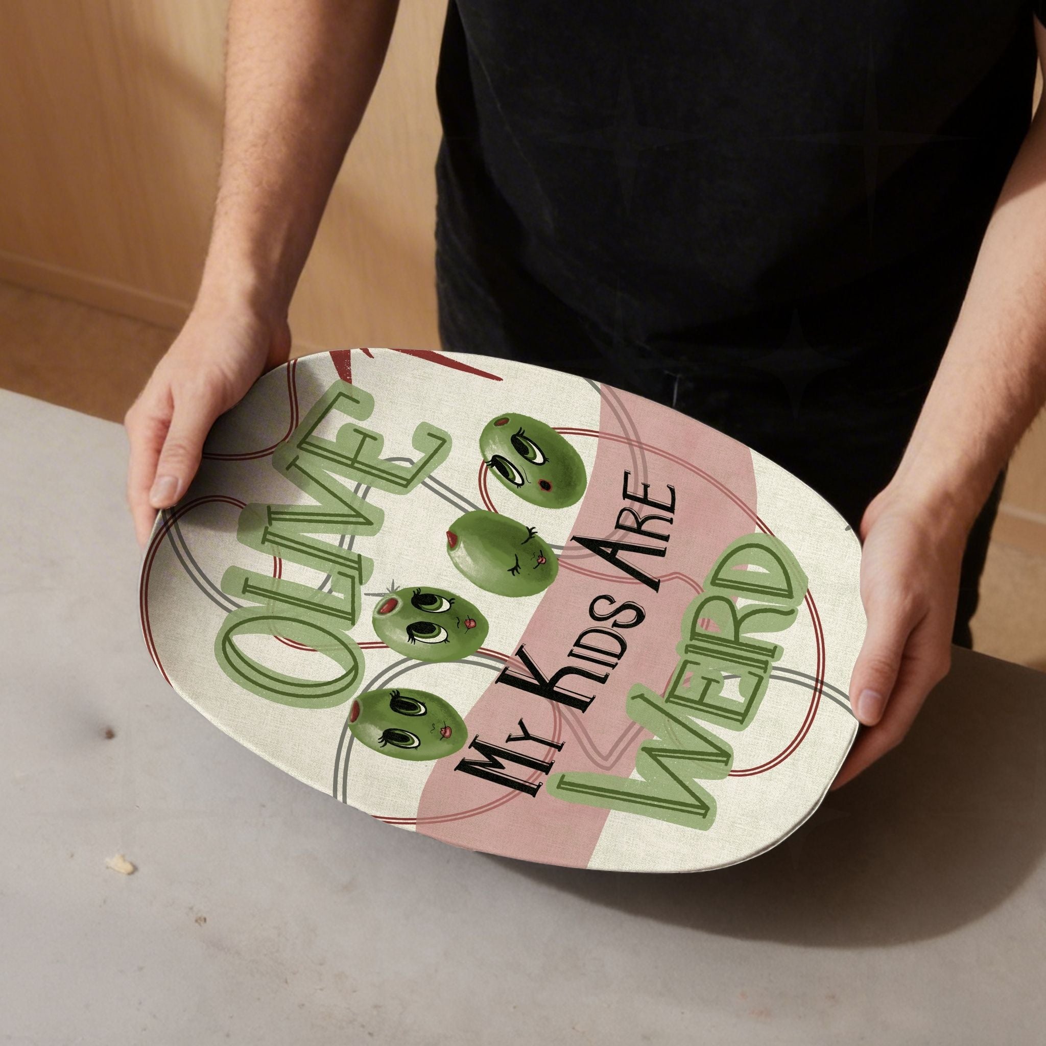 Man in black t-shirt holding funny retro serving platter with four cartoon olive characters and Olive My Kids Are Weird mid-century typography against warm wood background