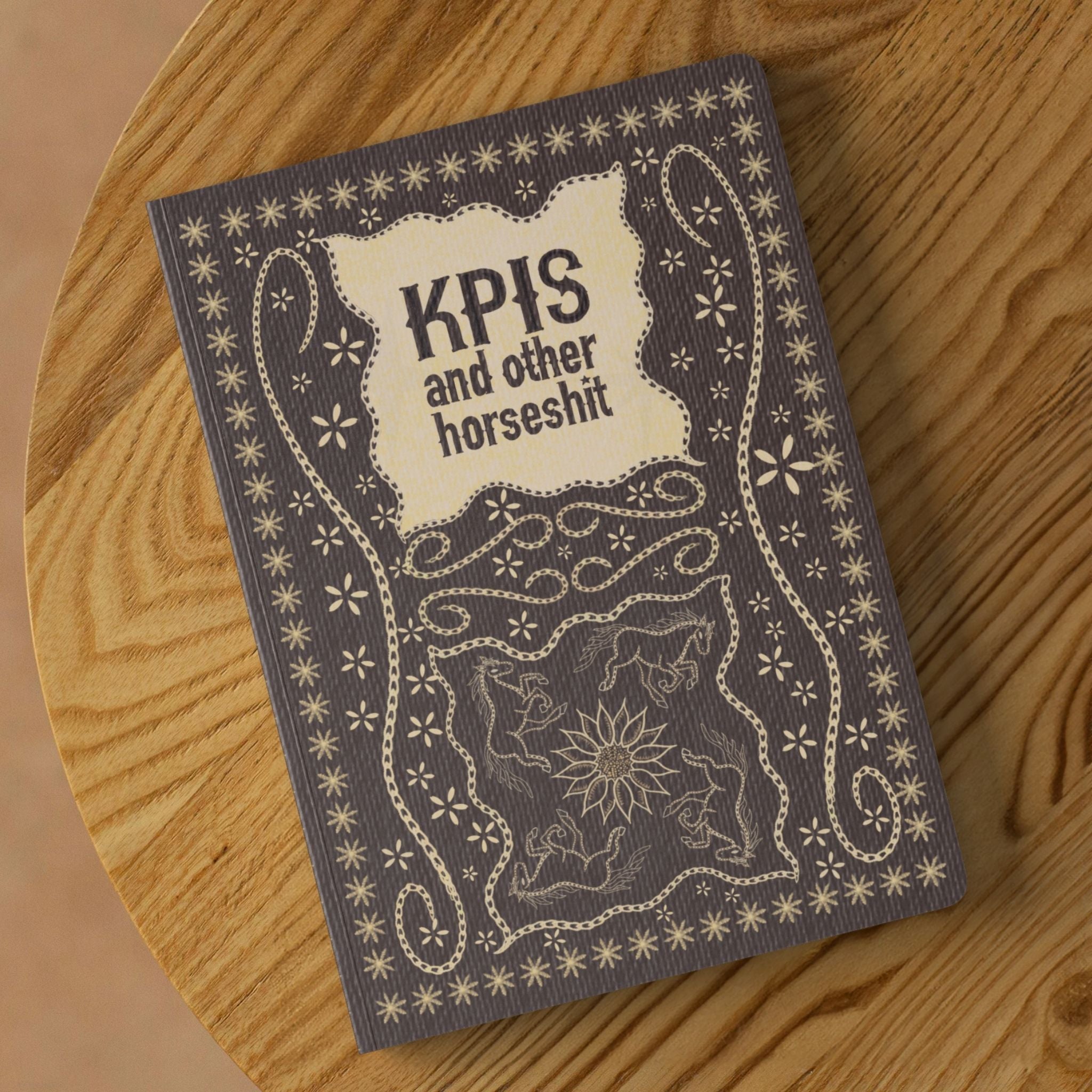Funny work journal with hand-stitched embroidery design reading KPIs and other horseshit on warm taupe softcover, displayed on round wooden surface showing full equestrian illustration