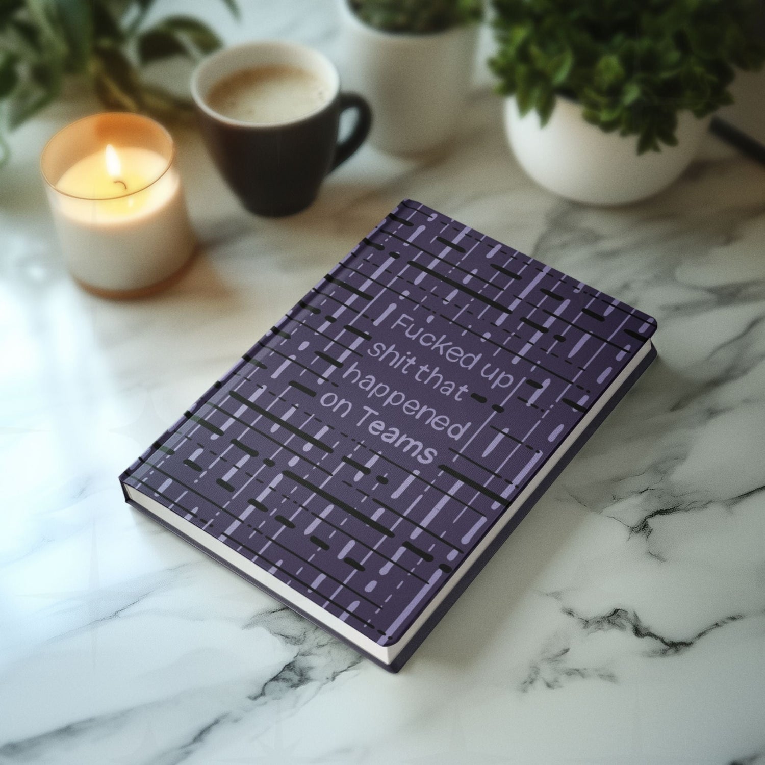 Funny Teams profanity hardcover journal lying on marble countertop next to lit candle, coffee mug, and green plant
