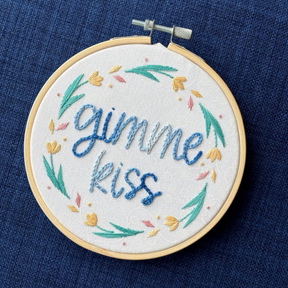 "Gimme kiss". Quote hand embroidered in handwriting font in variegated blues. Surrounding the text is a floral wreath with pastel yellow flowers, green stems and leaves, and yellow and pink floating petals. Sewn on white-on-white cotton. Framed as wall art in a light wood embroidery hoop.