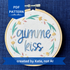 PDF pattern created by Kate, not AI. "Gimme kiss". Quote hand embroidered in handwriting font in variegated blues. Surrounding the text is a floral wreath with pastel yellow flowers, green stems and leaves, and yellow and pink floating petals. Sewn on white-on-white cotton. Framed as wall art in a light wood embroidery hoop.
