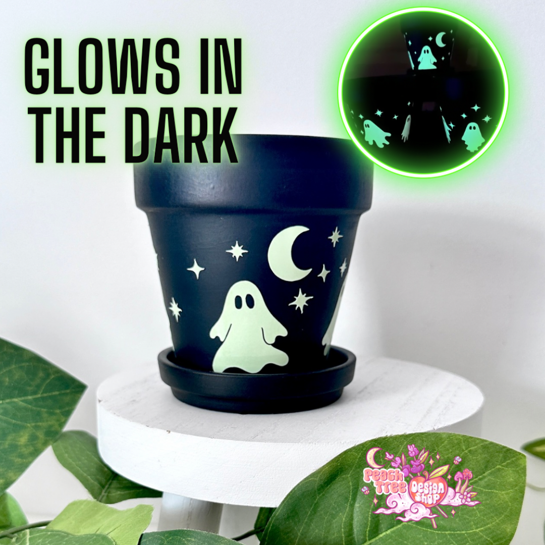 A Black 4" planter pot with a matching saucer decorated with glowing green ghost, moon, and star designs, displayed on a white pedestal with decorative leaves. Text on the image reads &