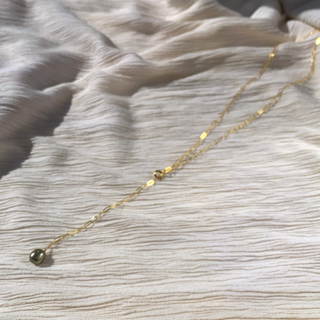 A gold-filled paperclip chain lariat necklace with a green fresh water pearl at the end, displayed on a textured white background.