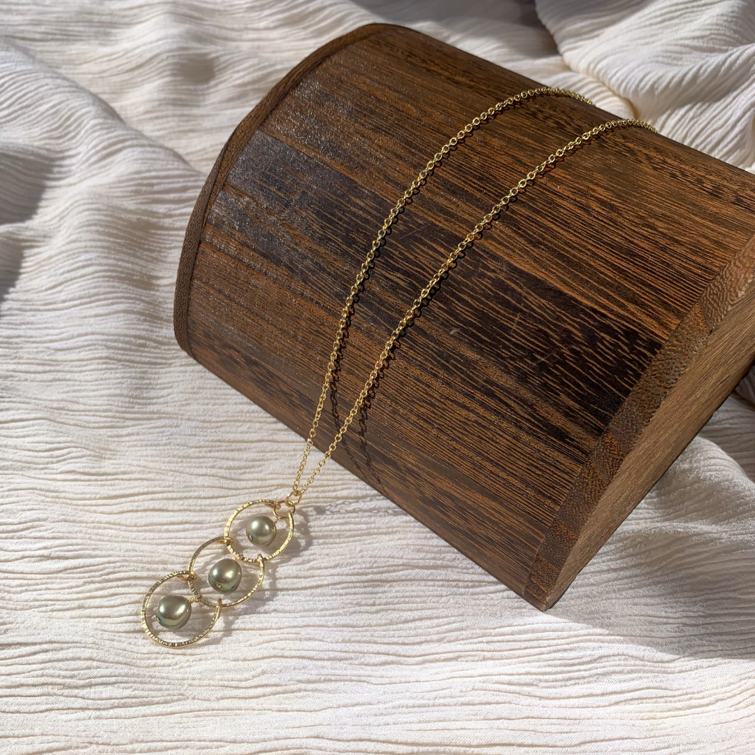 A gold-filled 14k necklace with three green fresh water pearls on a textured chain, displayed against a soft-focus background.