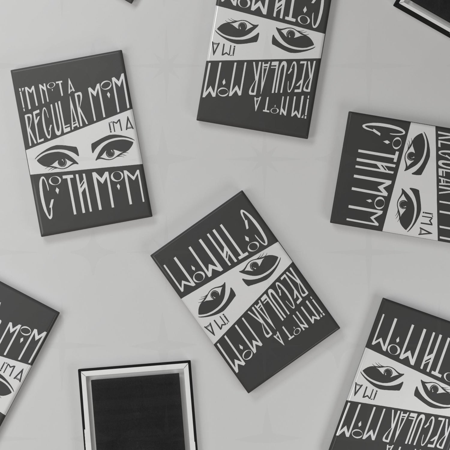 Flat lay of multiple goth mom magnets scattered at various angles on a light grey surface, showing both front design and black magnetic back