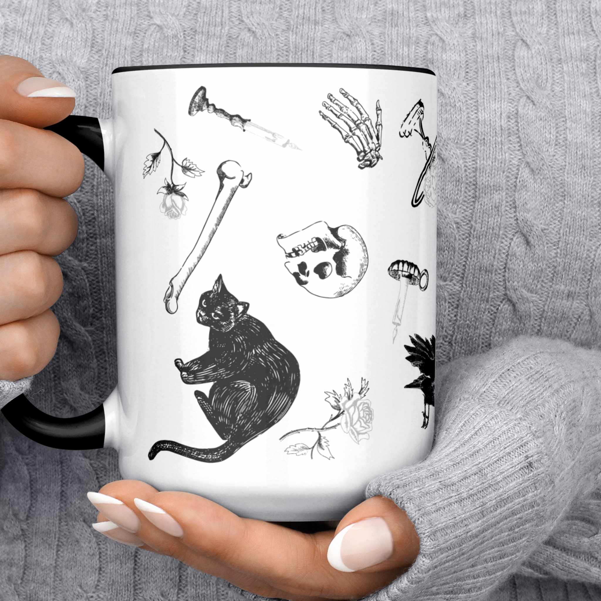 Black and white gothic pattern mug held in two hands showing black cats, a skull, and loose bones