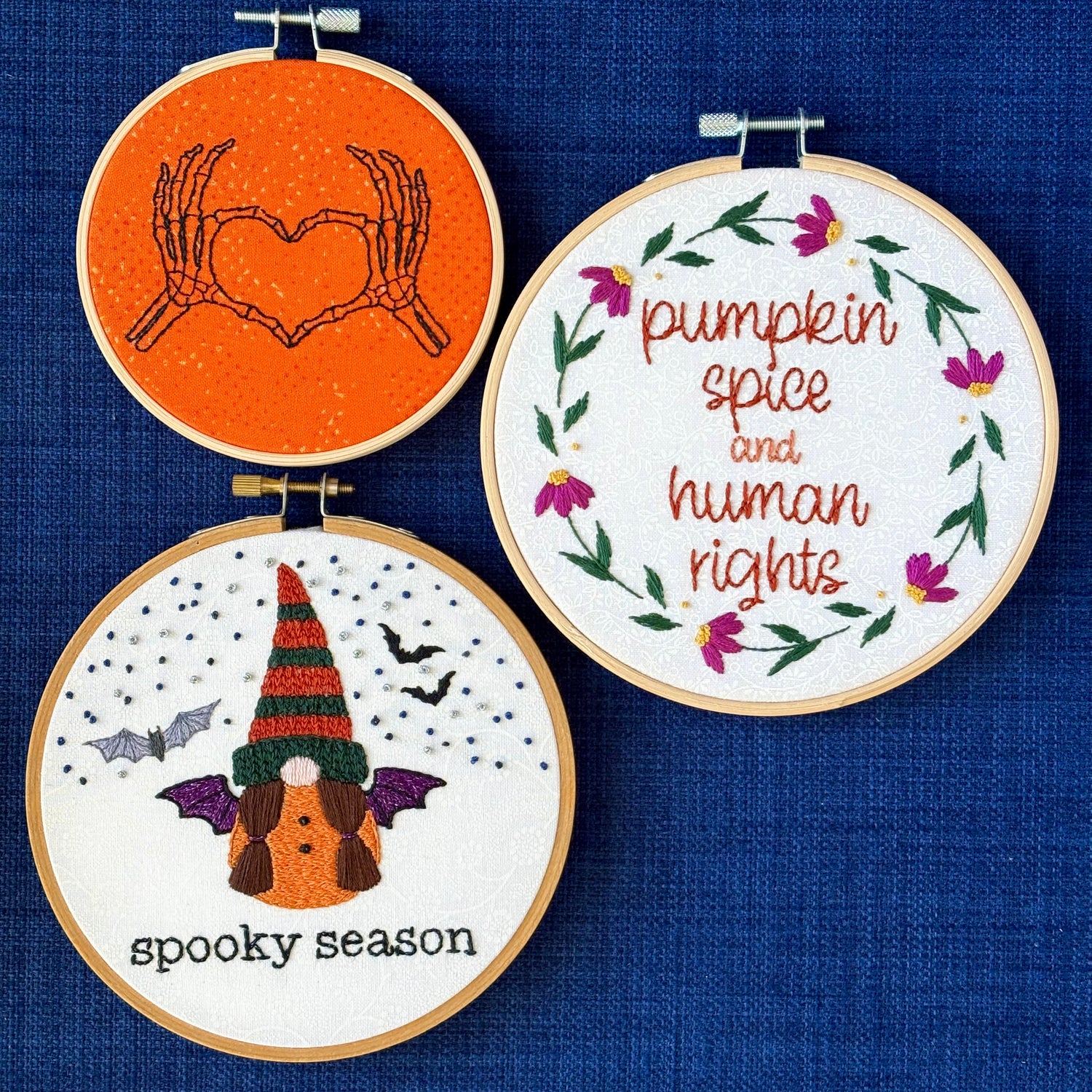 Hand embroidered pair of skeleton hands making a heart by touching thumbs and forefingers, while remaining fingers fan out. "Pumpkin spice and human rights". Handwriting font text surrounded by a floral wreath of pink asters and green leaves and stems. Hand embroidered gnome wears a an orange robe with black and sparkly purple bat wings. Under the gnome is "spooky season" embroidered in sparkling black thread. 3 designs displayed in light wooden hoops as wall art or Christmas ornaments.