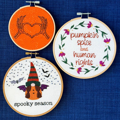 Hand embroidered pair of skeleton hands making a heart by touching thumbs and forefingers, while remaining fingers fan out. "Pumpkin spice and human rights". Handwriting font text surrounded by a floral wreath of pink asters and green leaves and stems. Hand embroidered gnome wears a an orange robe with black and sparkly purple bat wings. Under the gnome is "spooky season" embroidered in sparkling black thread. 3 designs displayed in light wooden hoops as wall art or Christmas ornaments.
