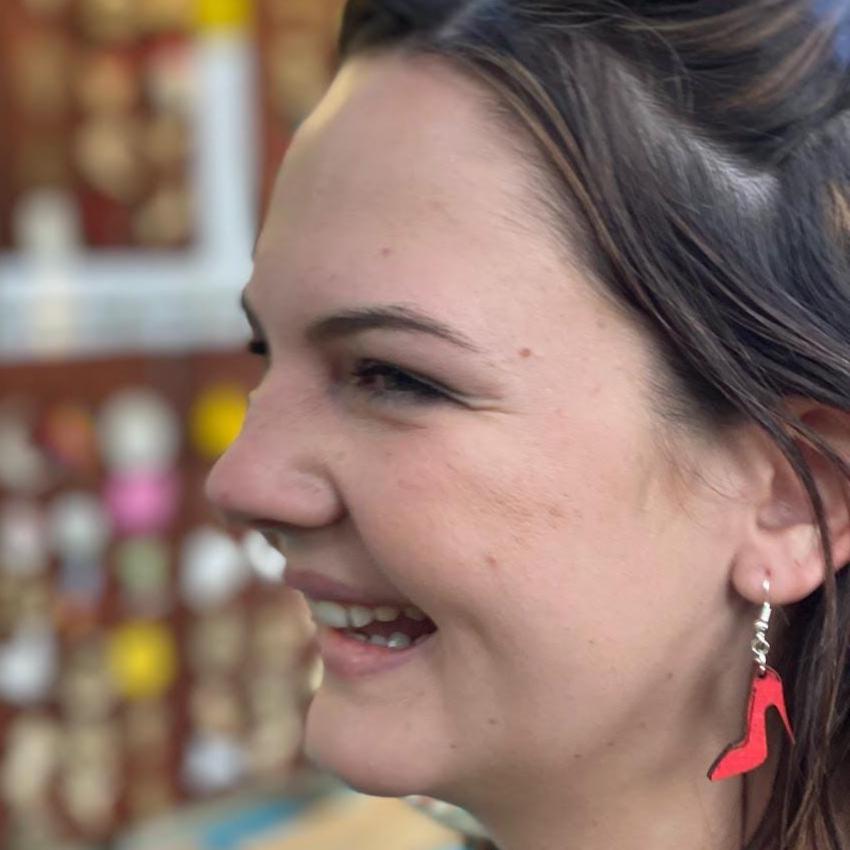 Person wearing hand-painted red high heel wooden dangle earring on a hypoallergenic .925 sterling silver hook, showing the lightweight size and fit when worn. Lightweight fashion jewelry inspired by high heels, perfect for fashion enthusiasts and stylish everyday wear.