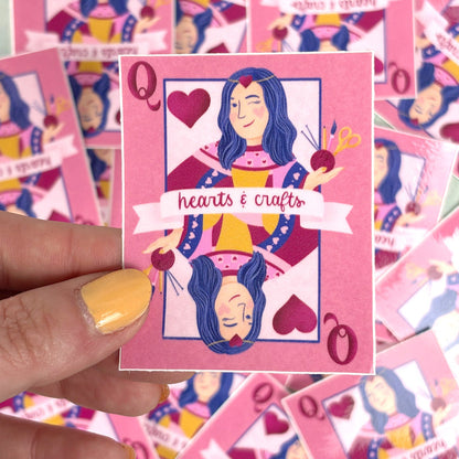 hand holding A pink sticker with a design of a queen playing card surrounded by hearts and crafting tools. The text &