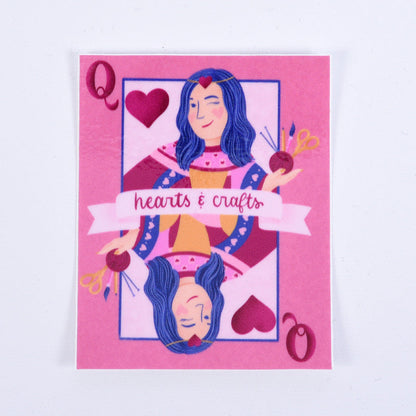 A pink sticker with a design of a queen playing card surrounded by hearts and crafting tools. The text &