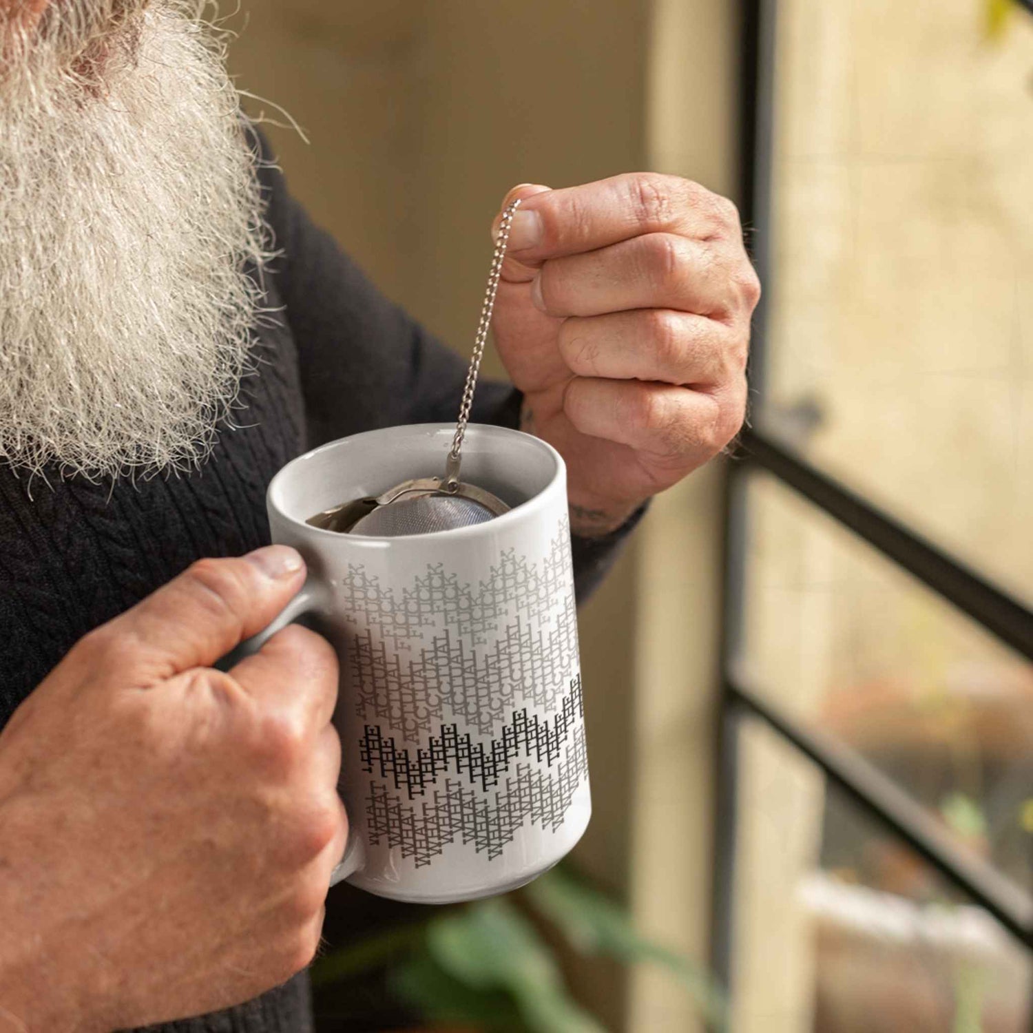  Person steeping tea with a metal infuser in a gray hidden profanity mug by a window, relaxed lifestyle setting