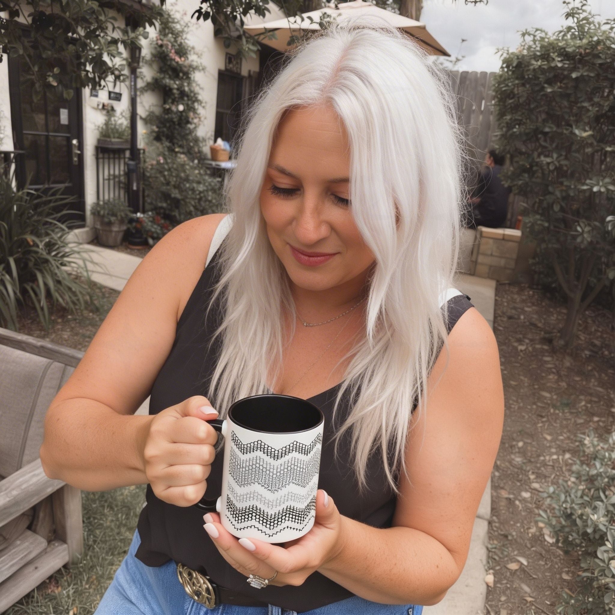 Lifestyle photo of a woman with white hair holding the Gray and Black hidden message mug with both hands at an outdoor garden cafe, looking down at the mug with a smile